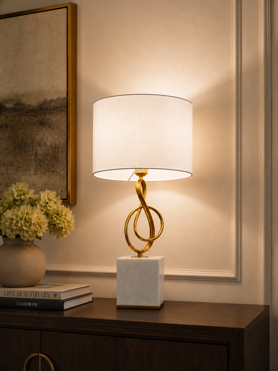 A table lamp with a white cylindrical shade, a gold twisted metal base, and a white marble square pedestal, placed on a dark wood console table in a warmly lit interior setting.