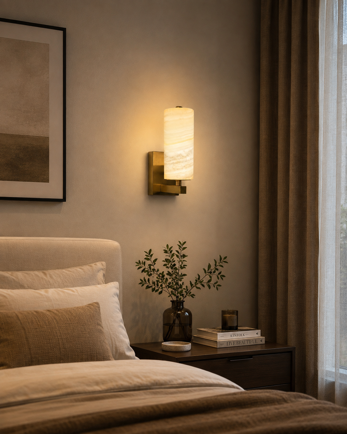 A modern wall sconce with a cylindrical white stone or marble shade and a brushed gold metal base, mounted on a beige wall in a styled bedroom setting next to a bed.