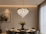 A modern dining room featuring a large circular crystal chandelier hanging over a round dining table with gray upholstered chairs, set against a warm beige wall with built-in lighting and sheer curtains.