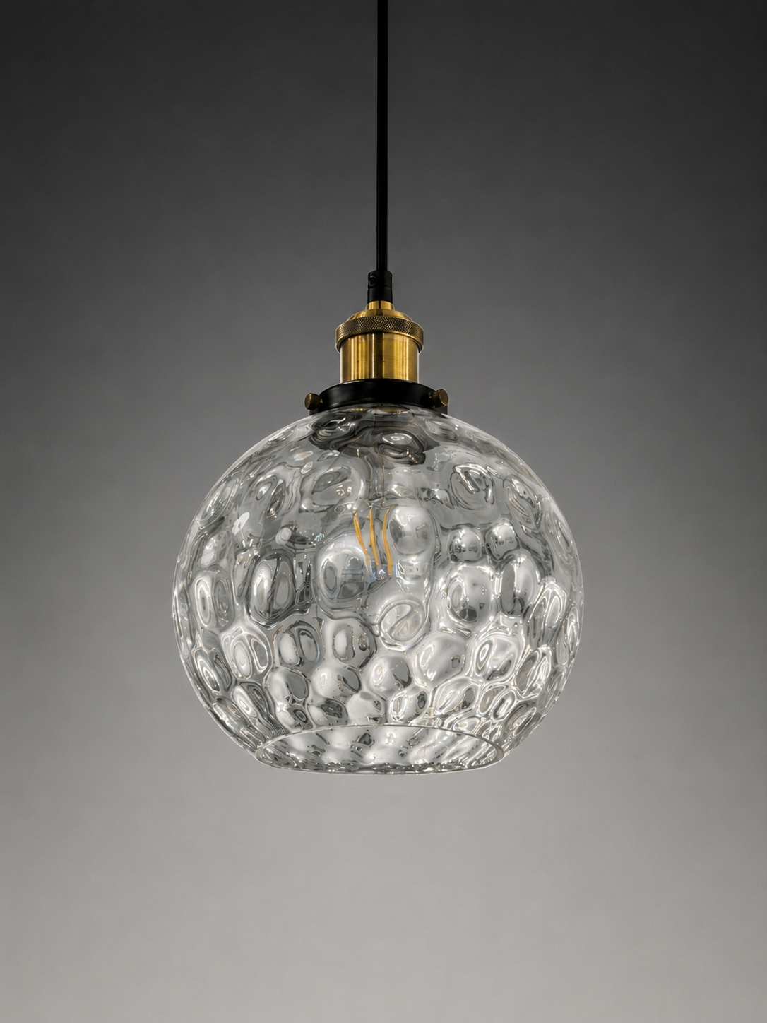 A clear textured glass pendant light with a hammered bubble pattern, suspended from a black cord with a gold metal cap, set against a dark gray gradient background.