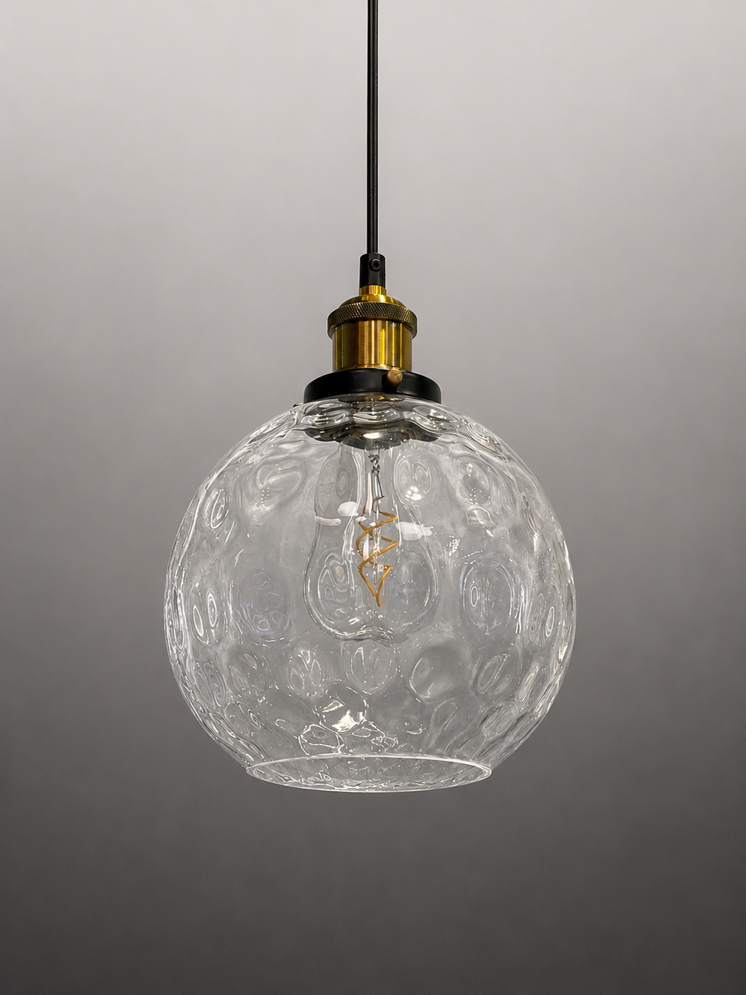 A clear textured glass globe pendant light with a black hanging cord and a brass-colored socket, shown against a neutral gray gradient background.