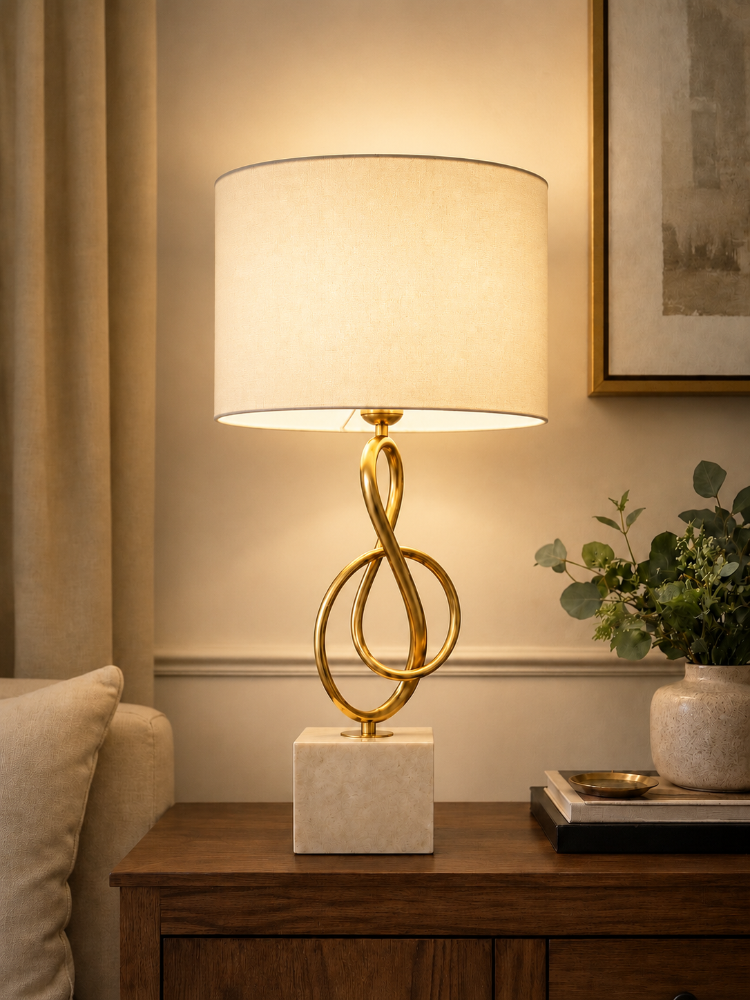A table lamp with a warm glowing drum shade, a gold-finished abstract twisted-ring metal base, and a square white marble-like pedestal, shown on a wooden side table in a styled interior setting.