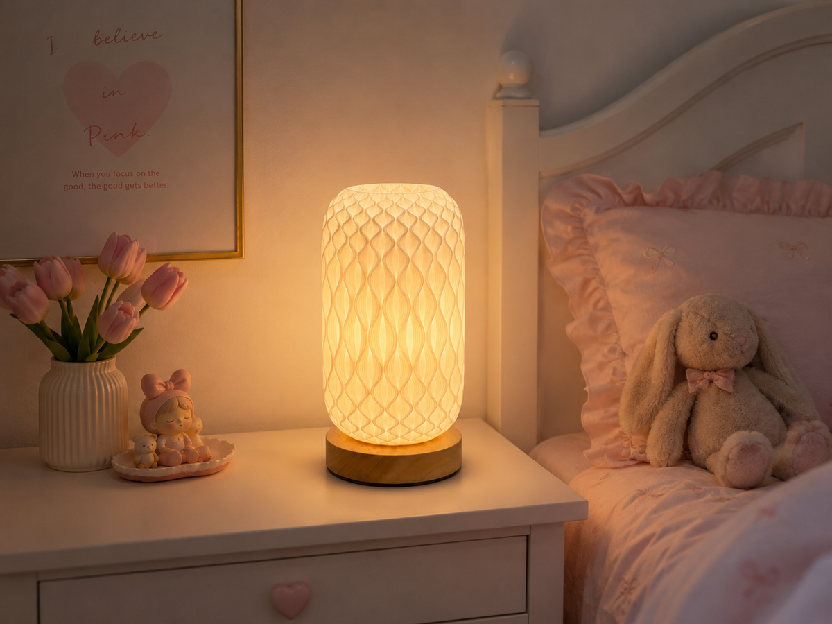 A glowing cylindrical table lamp with a textured honeycomb pattern and a gold-toned base sits on a white bedside table in a softly lit bedroom. Pink bedding, a stuffed bunny, tulips, and a framed print are visible in the background.