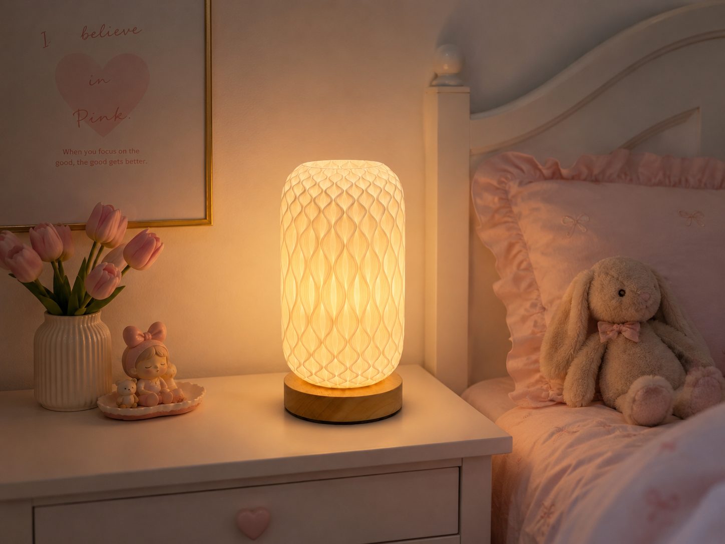 A glowing cylindrical table lamp with a textured honeycomb pattern and a gold-toned base sits on a white bedside table in a softly lit bedroom. Pink bedding, a stuffed bunny, tulips, and a framed print are visible in the background.
