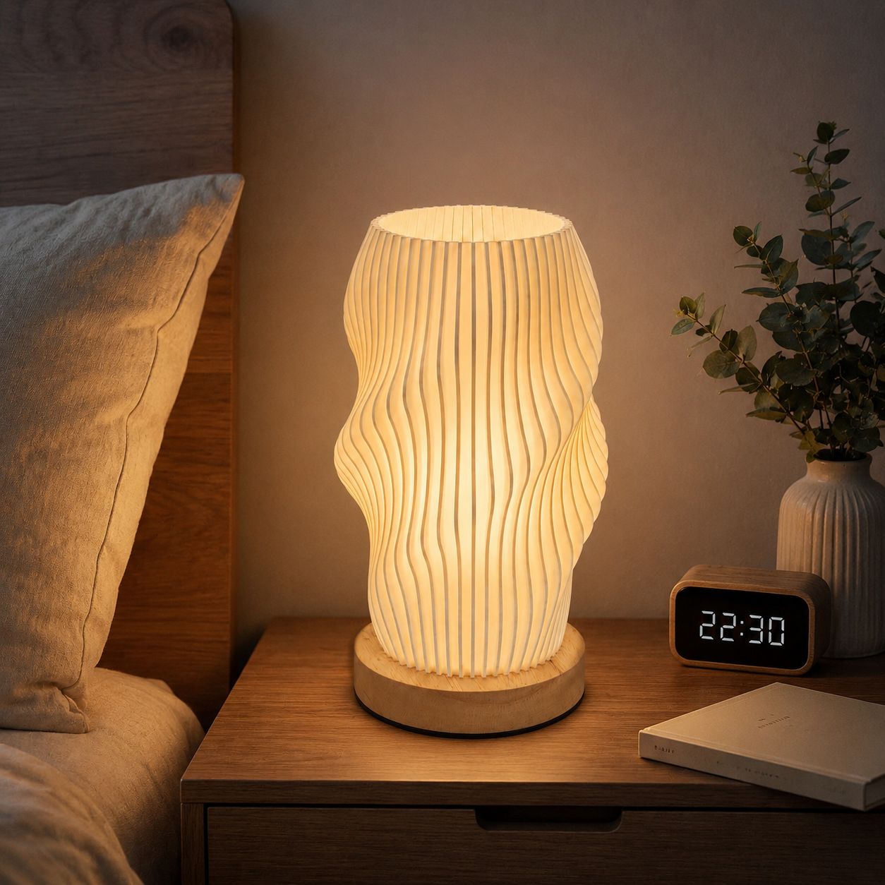 A modern wavy ribbed table lamp with a warm glowing light, sitting on a wooden bedside table next to a digital alarm clock, a book, and a potted plant.