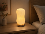 A glowing bedside table lamp with a ribbed, wavy translucent shade and a round wooden base, placed on a wooden nightstand next to a bed with a pillow and a small vase with greenery.