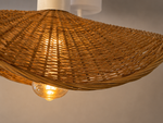 A close-up of a woven natural rattan pendant light shade glowing with warm light from an exposed bulb, against a neutral beige wall.